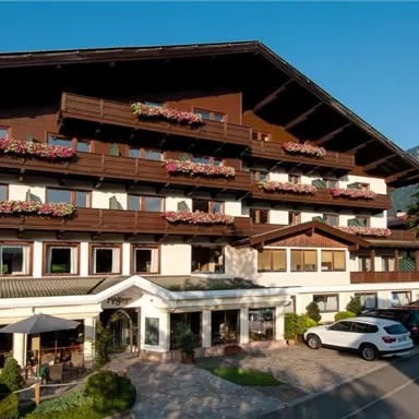 A cozy hotel in alpine style with balconies full of flowers. In the background, green mountains and a clear sky are visible.