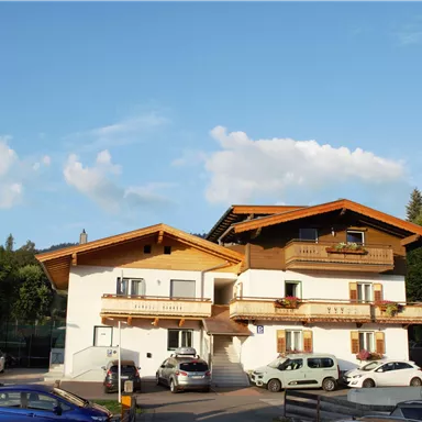 Ein komfortables Gebäude im alpinen Stil mit Holzverkleidungen und Balkonen. Vor dem Haus parken mehrere Autos unter einem klaren, blauen Himmel.