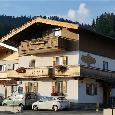 Ein traditionelles alpines Haus mit zwei Stockwerken und Holzveranda. Vor dem Haus stehen mehrere Autos geparkt.