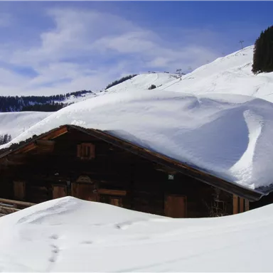 Ein gemütliches Holzhaus ist von tiefem Schnee umgeben. Im Hintergrund sieht man schneebedeckte Berge unter einem klaren blauen Himmel.