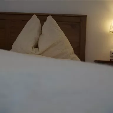 A cozy bedroom with a wooden bed and cushions. A soft lamp next to the bed creates a relaxed atmosphere.