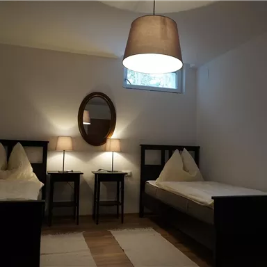 A bright bedroom with two single beds and a round mirror. Next to the beds are two nightstands with lamps.