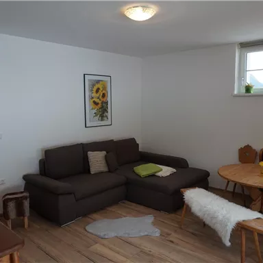 A cozy living room with a brown sofa and a round table. A picture of sunflowers hangs on the wall, and a window lets in plenty of daylight.