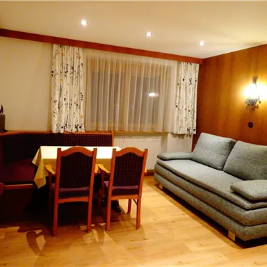 A cozy living room with a gray sofa and a dining table. The walls are made of wood, and there are large windows with curtains.