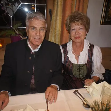 A couple in traditional attire is sitting at a laid table. In the background, decorative curtains and a warmly lit atmosphere can be seen.