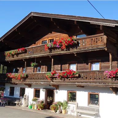 A traditional wooden house with blooming balcony flowers. In the background, trees and mountains are visible.