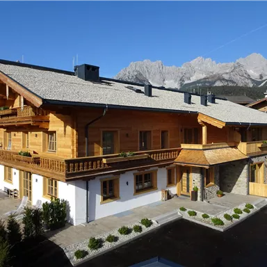 Ein schönes Holzhaus in einer malerischen Berglandschaft. Die Umgebung ist grün mit gepflegten Pflanzen und Blick auf die Berge im Hintergrund.