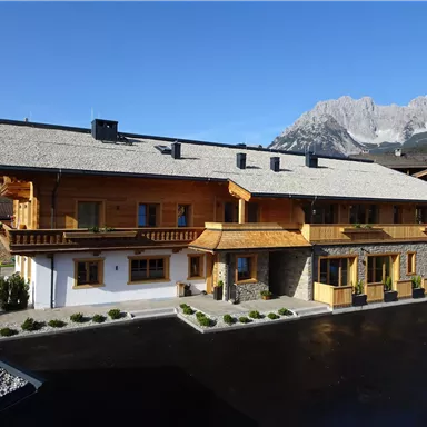 Ein wunderschönes Holzhaus in den Alpen mit einem steilen Dach. Im Hintergrund sind beeindruckende Berge und ein klarer blauer Himmel zu sehen.