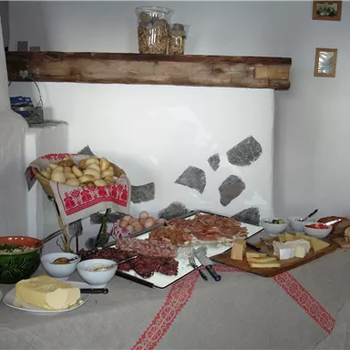 Ein gedeckter Tisch mit verschiedenen Käsesorten, Wurst und frischem Brot. Im Hintergrund sind Holzregale und Bilder an der Wand zu sehen.