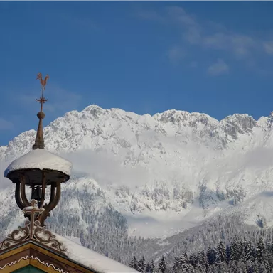 A snowy mountain landscape with high peaks and a clear sky. In the foreground, there is a traditional roof with a weather vane.