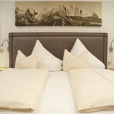 A cozy bedroom with a large bed and several pillows. On the wall hangs a picture of mountains.