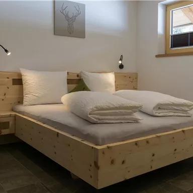 Ein gemütliches Schlafzimmer mit einem Holzbett und zwei großen, weißen Kissen. Es gibt sanfte Beleuchtung und ein Fenster mit Sicht auf die Außenwelt.