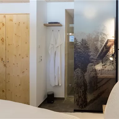 Ein gemütliches Schlafzimmer mit einem Holzschrank und einem modernen Badezimmer. Die Wände sind hell, während die Glastür ein schönes Bergmotiv zeigt.