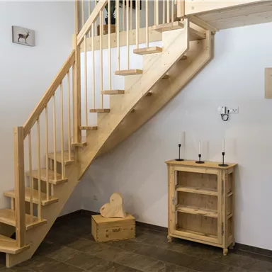 Ein heller Eingangsbereich mit einer Holztreppe und einem Holzschrank. Die Wände sind schlicht und leicht gestaltet.