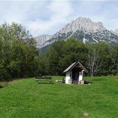 A picturesque landscape with a small house and a bench on a meadow. In the background, majestic mountains rise into the sky.