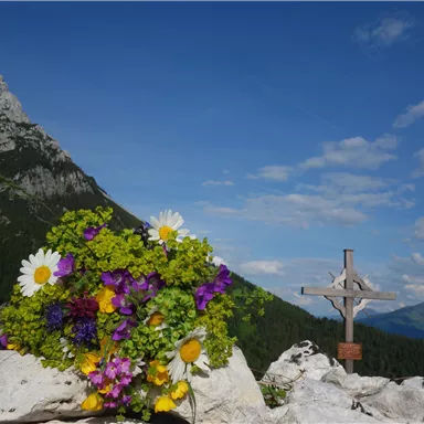 A bouquet of colorful herbs and flowers lies on a stone. In the background, majestic mountains and a cross can be seen.