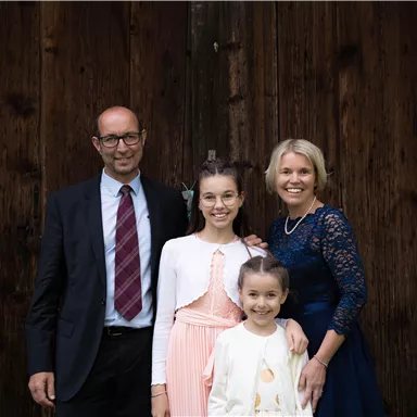 A family is standing in front of a wooden wall. They are smiling warmly and wearing festive clothing.