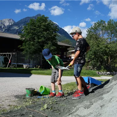 Two children are playing outside and digging in a pile of sand. In the background, trees and mountains can be seen.