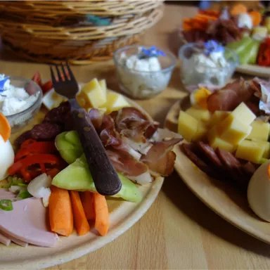 An appealingly arranged plate with various types of cold cuts, cheese, fresh vegetables, and small decorative eggs shaped like birds. Next to it are small bowls with dip and other side dishes.