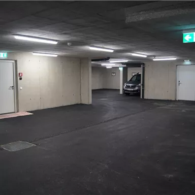 A modern parking garage area with gray walls and bright light. In the background, there is a car in front of an open door.