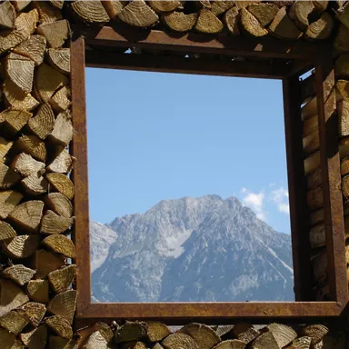 A wooden log window frame offers a view of a majestic mountain and the blue sky. The scene conveys tranquility and closeness to nature.