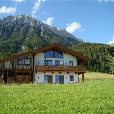 Ein charmantes Haus im bayerischen Stil, umgeben von grünen Wiesen und majestätischen Bergen. Der klare Himmel und die Natur schaffen eine ruhige Atmosphäre.