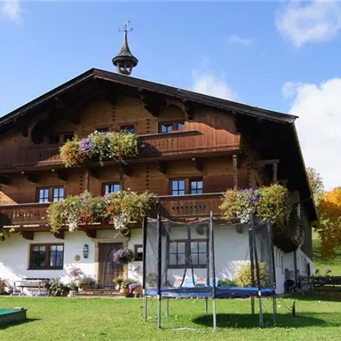 Ein traditionelles, hölzernes Bauernhaus mit vielen Balkonblumen. Im Vordergrund befindet sich ein Trampolin und bunte Bäume im Hintergrund.