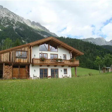 Ein gemütliches Haus im alpenländischen Stil, umgeben von Wiesen und Bergen. Die Landschaft ist grün und lädt zum Entspannen ein.