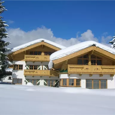 Ein hübsches Chalet im Schnee mit einem großen Dach aus Holz. Es ist umgeben von schneebedeckten Bäumen und einem klaren Himmel.