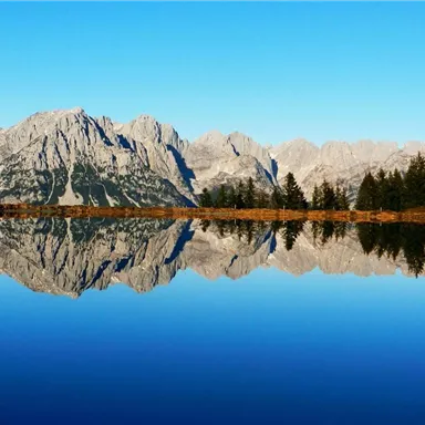 A picturesque mountain landscape with clear waters reflecting the peaks. The sky is bright blue and conveys a tranquil atmosphere.