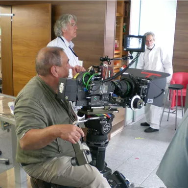 A cameraman sits in front of a large camera. In the background, there are additional people and a modern room ambiance.