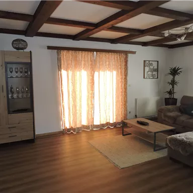 Ein gemütliches Wohnzimmer mit Holzdecke, einem Sofa und einem Couchtisch. Große Fenster mit Vorhängen sorgen für helles Licht und eine einladende Atmosphäre.