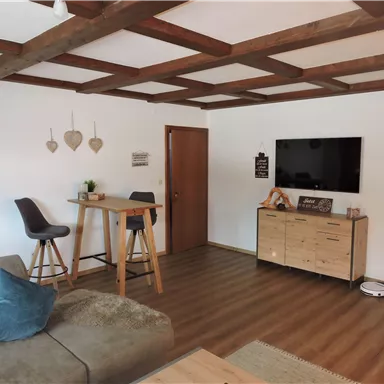 Ein helles Wohnzimmer mit Holzbalkendecke und stylischer Einrichtung. Im Raum stehen ein Sofa, ein Esstisch und ein Fernseher an der Wand.