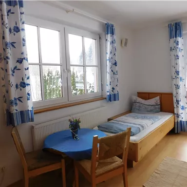 A bright room with a single bed and a table. Large windows let in a lot of light.