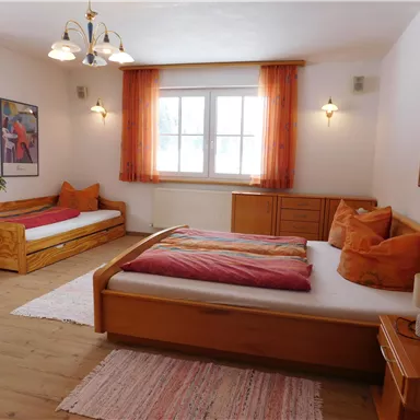 A bright bedroom with two beds, a large window, and orange curtains. The room features wooden furniture and a cozy atmosphere.