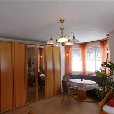 A bright room with a large mirror wardrobe and a round table. The windows are adorned with orange curtains, and there are plants in the room.