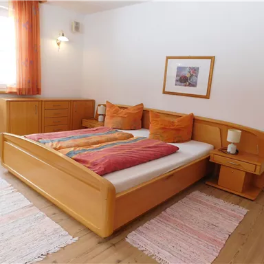 A cozy bedroom with a double bed and wooden furniture. The windows are decorated with orange curtains and there is a bedside table with a lamp.