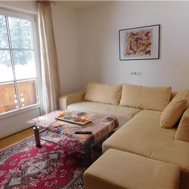 A cozy living room with a beige sofa and a small table. In the background, there is a window and a balcony door that offer a view of the snow.