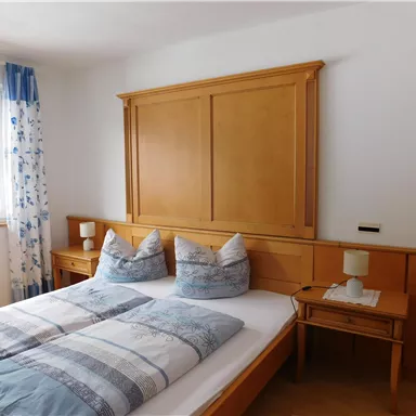 A bright bedroom with a double bed and wooden paneling. The windows are decorated with blue curtains, and a nightstand stands next to the bed.