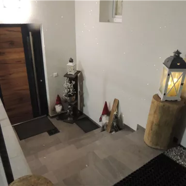 Ein Eingangsbereich mit einer modernen Tür, einem Steinboden und einer Laterne. Dekorative Elemente wie kleine Figuren und Holzstämme verleihen dem Raum eine gemütliche Atmosphäre.