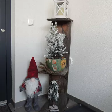 Eine festliche Ecke mit einem Holzregal, das einen kleinen, mit Schnee bedeckten Baum und eine Laterne zeigt. Neben dem Regal steht eine große Zwergfigur in roten und grauen Farben.