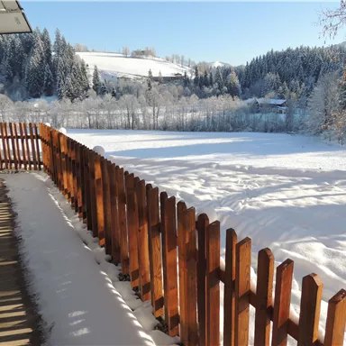 Ein verschneiter Winterlandschaft mit einem hölzernen Zaun. Im Hintergrund sind schneebedeckte Bäume und sanfte Hügel sichtbar.