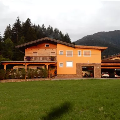 Ein orangefarbenes Haus mit einem großen Garten und einer angrenzenden Garage. Im Hintergrund sind Berge und Bäume sichtbar.