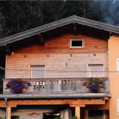 Ein gemütliches Haus mit einem Holzbalcon und bunten Blumen. Im Hintergrund sind hohe, grüne Bäume zu sehen.