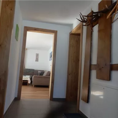 Ein heller Flur mit Holzdetails und einem Sichtfenster zum Wohnzimmer. Auf der linken Seite hängen Kleiderhaken an der Wand.