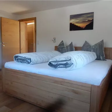 Ein schlichtes Schlafzimmer mit einem großen Holzbett und zwei Kissen. Die Wände sind hell und eine Tür führt zu einem weiteren Raum.