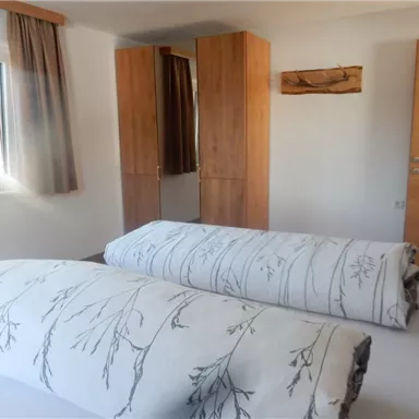 Ein helles Schlafzimmer mit einem Fenster und einem Kleiderschrank aus Holz. Auf dem Bett liegen dicke Decken mit einem floralen Muster.