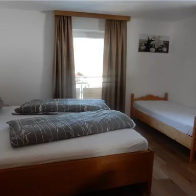 Ein einfaches Schlafzimmer mit zwei Betten und einem Einzelbett. Das Zimmer hat große Fenster und helle Vorhänge.