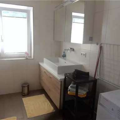 Ein modernes Badezimmer mit einer weißen Waschtischanlage und einem großen Spiegel. Es gibt ein Fenster, eine Waschmaschine und eine Wäscheablage neben bunten Handtüchern.