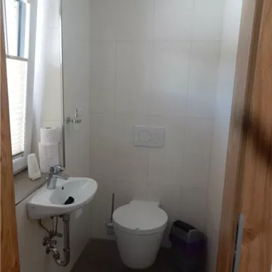 Ein kleines, modernes Badezimmer mit einer Toilette und einem Waschbecken. Es hat helle Wände und ein Fenster, das Tageslicht hereinlässt.